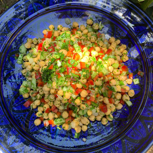 A prepared chickpea salad on a plate, featuring ingredients such as chickpeas, herbs, red capsicum, red onions, and celery, served with a separate bowl of dressing.