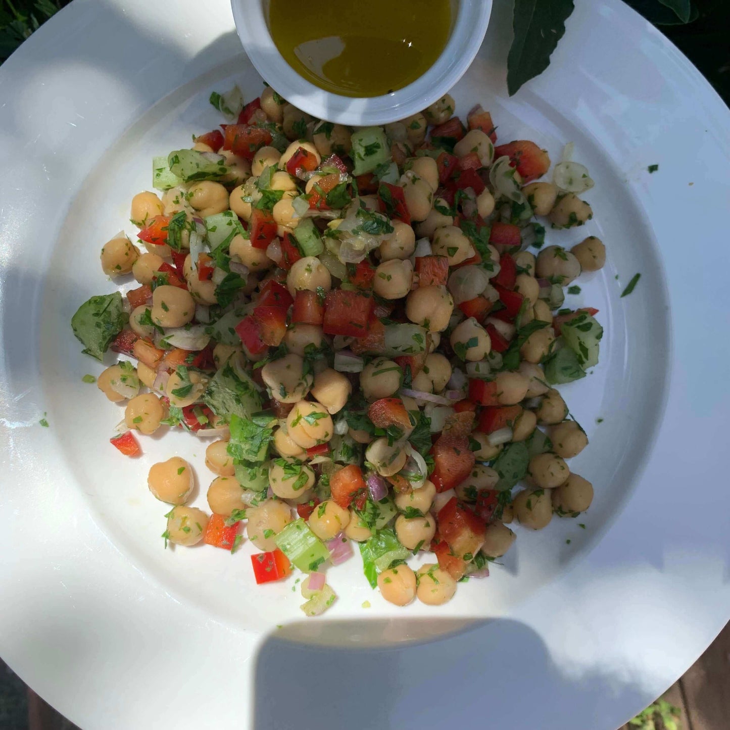 A prepared chickpea salad on a plate, featuring ingredients such as chickpeas, herbs, red capsicum, red onions, and celery, served with a separate bowl of dressing.